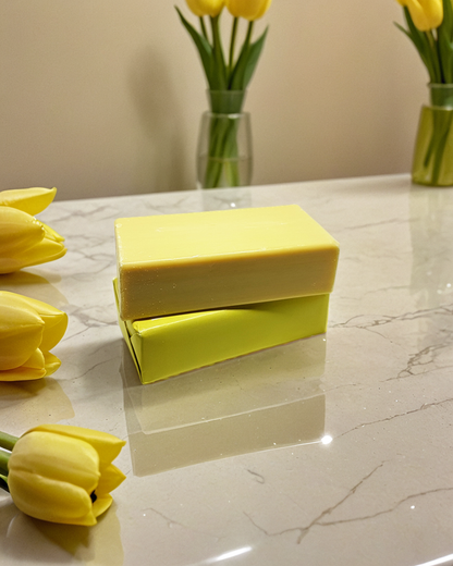 Yellow handmade soap bar displayed on a marble countertop, with yellow tulip flowers elegantly arranged around it and in vases in the background, creating a fresh, clean, and natural aesthetic suitable for a skincare or beauty product listing.